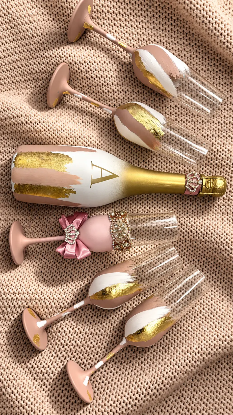 Juego de botella y copas en rosa blush, blanco y dorado sobre tejido beige